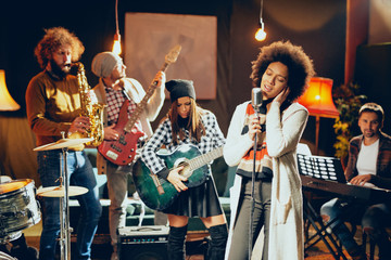 Mixed race woman singing. In background band playing instruments. Home studio interior. - obrazy, fototapety, plakaty