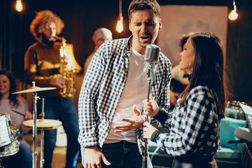 Singers practicing for duet. In background band playing. Home studio interior.