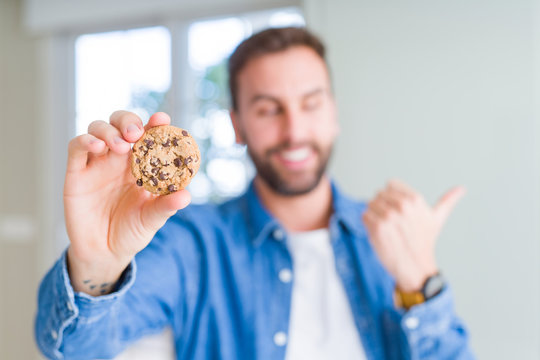 Handsome man eating chocolate chips cookies pointing and showing with thumb up to the side with happy face smiling