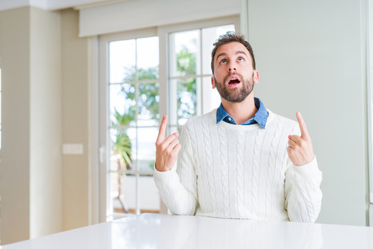 Handsome man wearing casual sweater amazed and surprised looking up and pointing with fingers and raised arms.