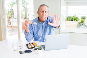 Handsome business senior man eating delivery sushi while working using laptop with open hand doing stop sign with serious and confident expression, defense gesture