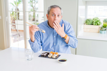 Handsome senior man eating take away sushi using chopsticks at home cover mouth with hand shocked with shame for mistake, expression of fear, scared in silence, secret concept