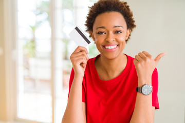 Young african american woman holding credit card pointing and showing with thumb up to the side with happy face smiling