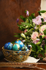 Painted colored Easter eggs in golden bowl and copper mug near bouquet of flowers on red animal skin. Boho stile.