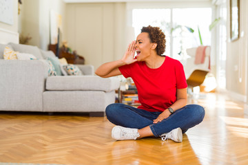 Young beautiful african american woman sitting on the floor at home shouting and screaming loud to side with hand on mouth. Communication concept.