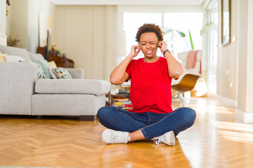 Young beautiful african american woman sitting on the floor at home covering ears with fingers with annoyed expression for the noise of loud music. Deaf concept.