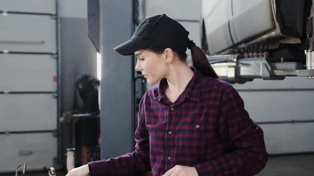 The girl-car mechanic, beautiful appearance, brunette, in a black cap and a plaid shirt, carries out repair work with a car on a lift at a service station