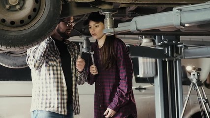 A beautiful girl, an auto mechanic, is learning how to repair a car's suspension from a more experienced man, an auto mechanic. The choice of tools, wrenches, for repairing the suspension of the car - Powered by Adobe
