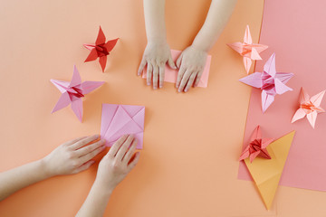 Children's hands do origami from colored paper on living coral background.