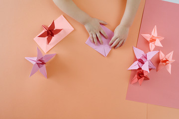 Children's hands do origami from colored paper on living coral background.
