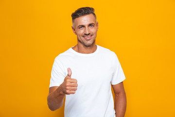 Handsome emotional man posing isolated over yellow wall background showing thumbs up gesture. - obrazy, fototapety, plakaty