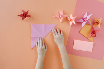 Children's hands do origami from colored paper on living coral background.