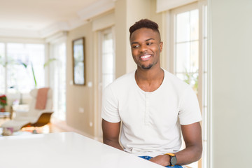 Handsome african american man on white table at home looking away to side with smile on face, natural expression. Laughing confident.