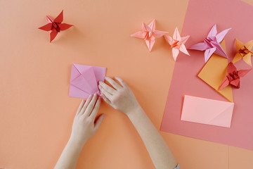 Children's hands do origami from colored paper on living coral background.