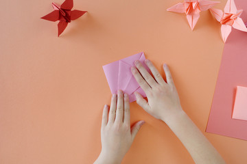 Children's hands do origami from colored paper on living coral background.