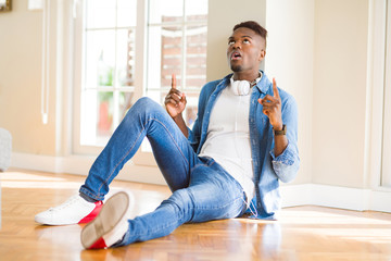 African american man wearing headphones listening to music sitting on the floor amazed and surprised looking up and pointing with fingers and raised arms.