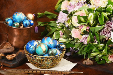 Painted colored Easter eggs in golden bowl and copper mug near bouquet of flowers on red animal...