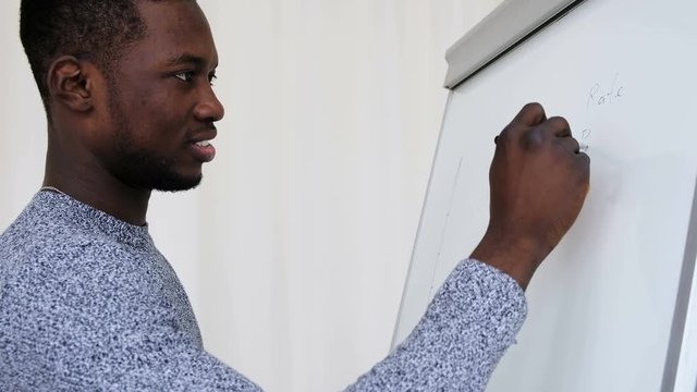 Side view young African male student of business school drawing profit diagram on whiteboard ready to lesson class work. Confident man writing on flip chart during studying low angle
