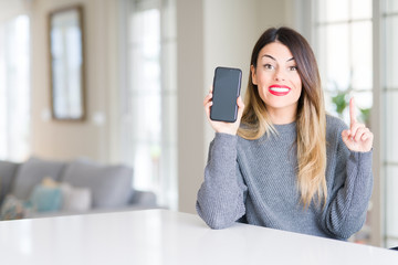 Young beautiful woman showing smartphone screen at home surprised with an idea or question pointing finger with happy face, number one