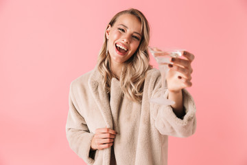 Emotional young pretty woman posing isolated over pink wall background holding glass with alcohol drink.