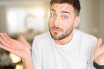 Young handsome man wearing casual white t-shirt at home clueless and confused expression with arms and hands raised. Doubt concept.