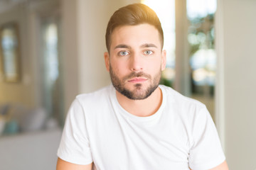 Obraz premium Young handsome man wearing casual white t-shirt at home with serious expression on face. Simple and natural looking at the camera.
