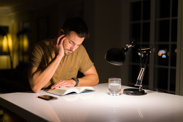 Young handsome man studying at home, reading a book at night