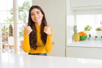 Young beautiful woman at home on white table success sign doing positive gesture with hand, thumbs up smiling and happy. Looking at the camera with cheerful expression, winner gesture.