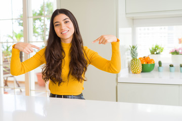 Young beautiful woman at home on white table looking confident with smile on face, pointing oneself...