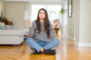 Young beautiful woman sitting on the floor at home clueless and confused expression with arms and hands raised. Doubt concept.