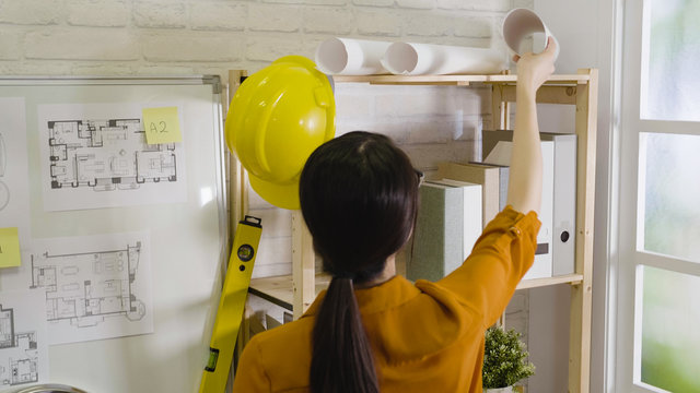 back view of asian girl architect standing high to take down blueprint paper on top bookshelf near white board with floor plan. woman interior designer ready work in office. hard hat hang on shelf