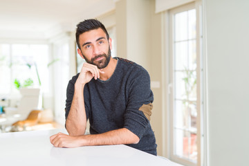 Handsome hispanic man wearing casual sweater at home with hand on chin thinking about question, pensive expression. Smiling with thoughtful face. Doubt concept.