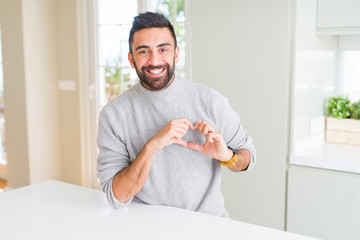 Handsome hispanic man wearing casual sweater at home smiling in love showing heart symbol and shape with hands. Romantic concept.