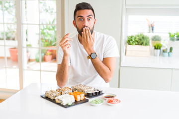 Handsome hispanic man eating asian sushi using chopsticks cover mouth with hand shocked with shame for mistake, expression of fear, scared in silence, secret concept