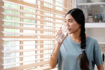Youn beautiful asian woman drinking water while standing by window in kitchen background, peolpe...