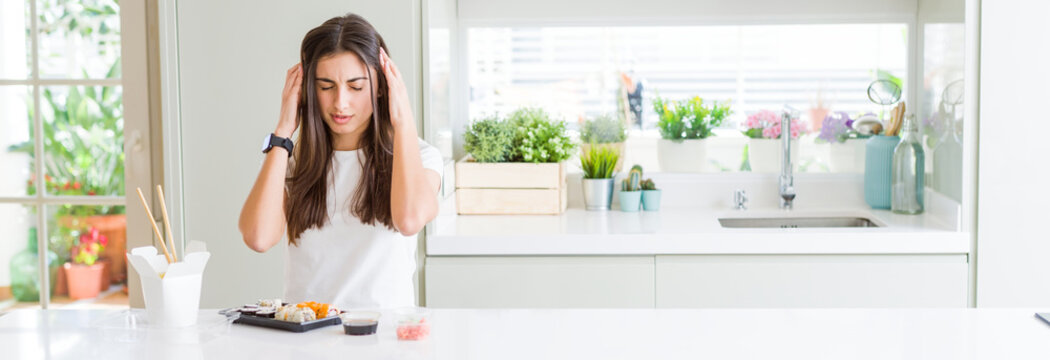 Wide angle picture of beautiful young woman eating asian sushi from delivery with hand on head for pain in head because stress. Suffering migraine.
