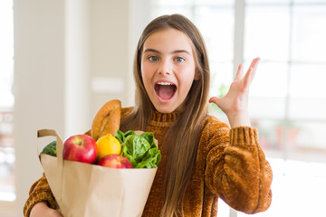 Beautiful young girl holding paper bag of fresh groceries very happy and excited, winner expression celebrating victory screaming with big smile and raised hands