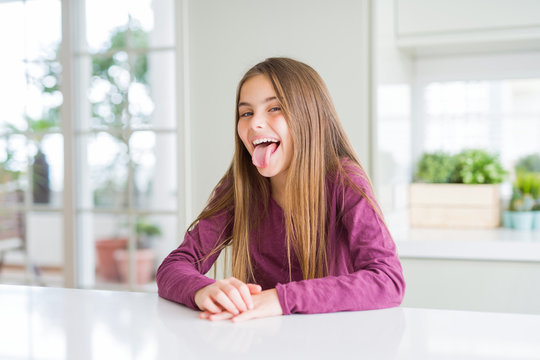 Beautiful young girl kid on white table sticking tongue out happy with funny expression. Emotion concept.