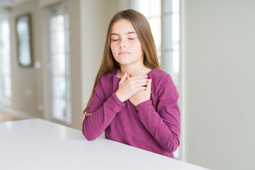 Beautiful young girl kid on white table smiling with hands on chest with closed eyes and grateful gesture on face. Health concept.