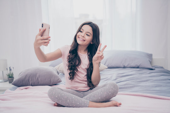 Close up photo cute beautiful she her little girl sitting bed smart phone hands video call show v-sign say hi relatives wear home t-shirt pants comfortable apartments flat bright light colored room