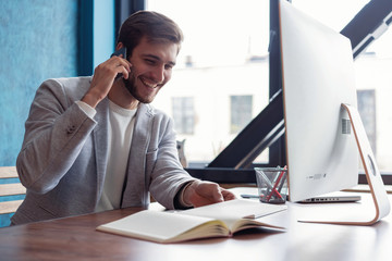 Fototapeta premium Young smiling bearded businessman is sitting at table in front of computer, talking on cell phone. Freelancer, entrepreneur works at coworking centre. Telephone conversations.