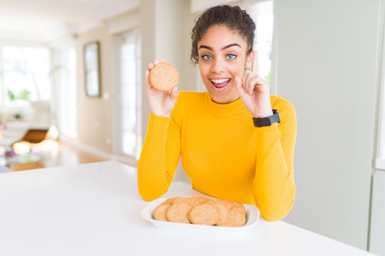 Young African American Girl Eating Healthy Whole Grain Biscuits Surprised With An Idea Or Question Pointing Finger With Happy Face, Number One