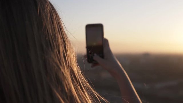 Long haired young woman standing from the back uses mobile phone at city blurred background with sunset or sunrise. Enjoying the time, making photo of her hand in front the sun rays while standing on