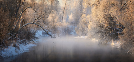 Winter landscape with river, snow-covered trees, fog and hoarfrost. Winter sunrise on river with fog.