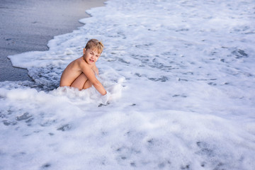 Little blonde child boy siting on sea shore and playing in waves