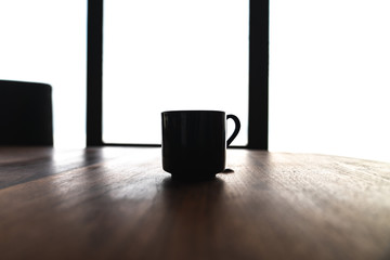 A single black porcelain or ceramic coffee cup or mug with hot tea sitting on a walnut wood colored dining table with bright daylight coming from windows beyond in a modern highrise condo building.