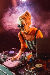 cheerful dj girl in sunglasses touching dj equipment in nightclub with smoke