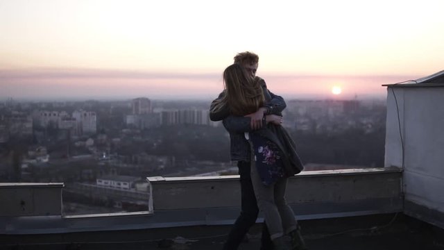 Redhead guy whirls his girlfriend on the roof with a cityscape and sunset horizon on the background. Happy time together, dating in romantic places
