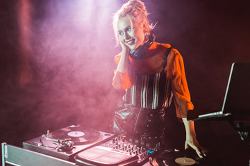 happy and stylish dj girl in headphones standing near dj mixer in nightclub with smoke