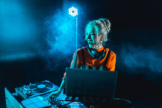 happy dj girl in headphones standing near dj mixer and laptop in nightclub with smoke